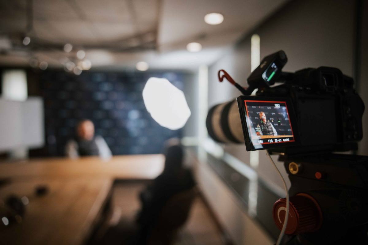 A cinema camera filming a speaker across the room during a recorded session.