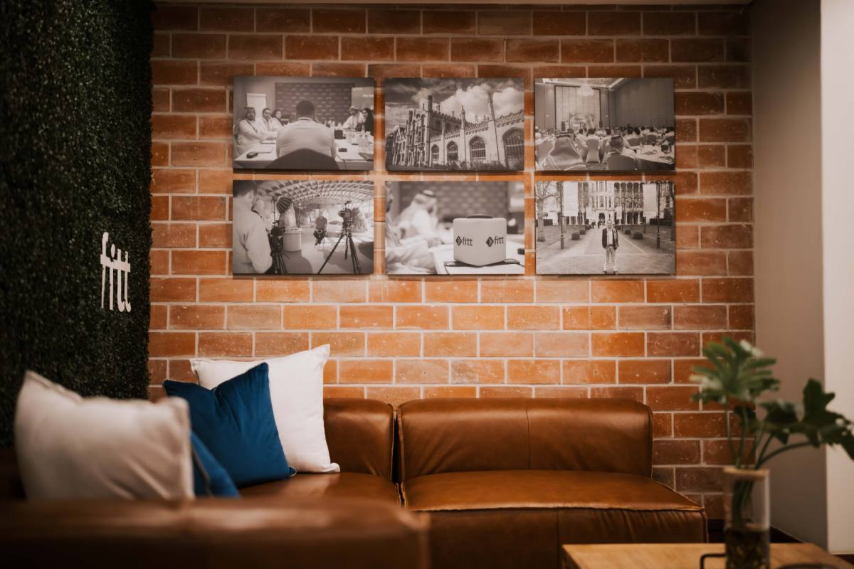 Lounge with a leather couch beneath a gallery of framed programme photography on an exposed brick wall.